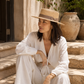 Woman in a white outfit and straw hat sitting on stone steps outdoors.