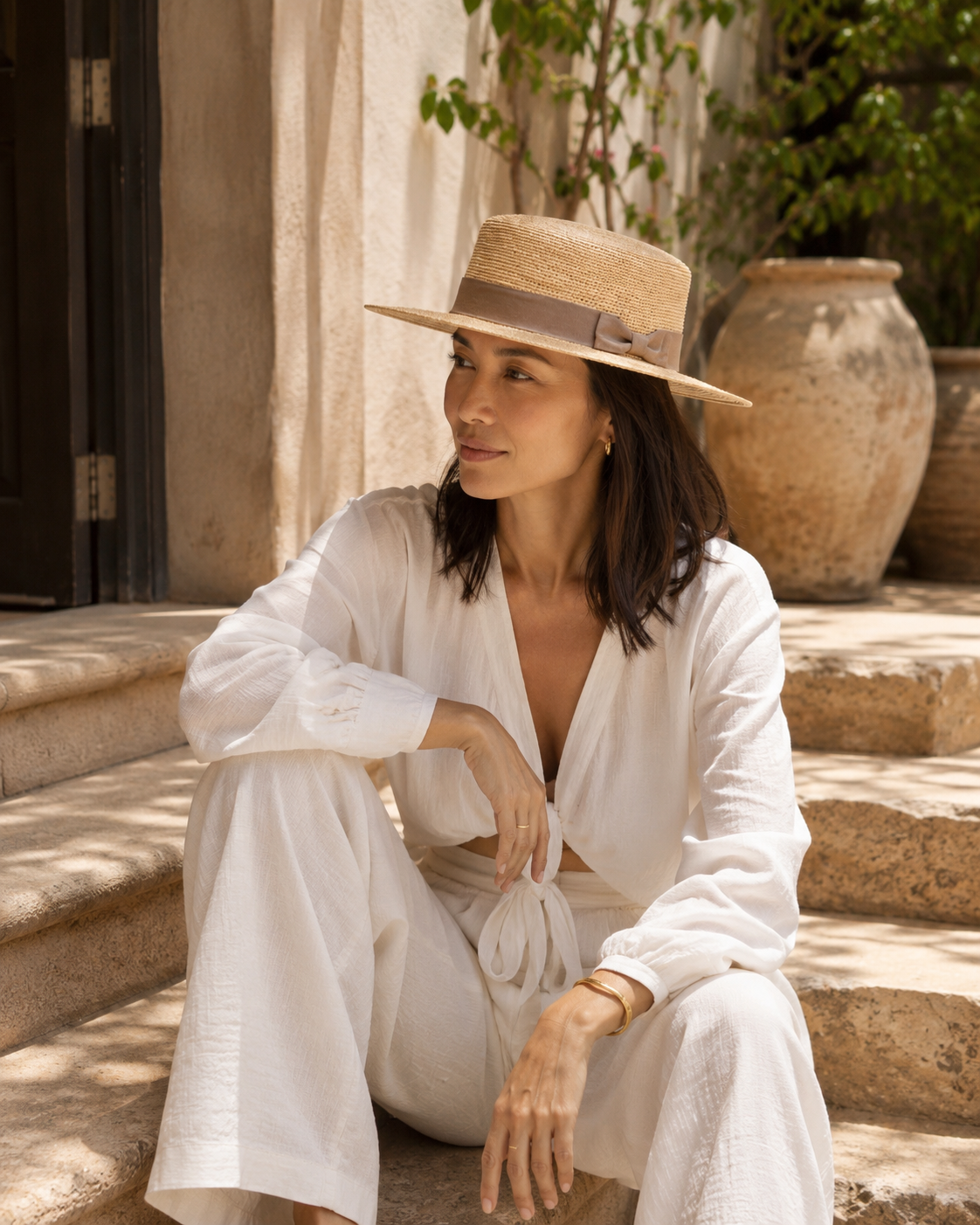 Woman in a white outfit and straw hat sitting on stone steps outdoors.