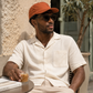 Man sitting outdoors wearing a beige shirt, orange cap, and sunglasses, holding a drink.