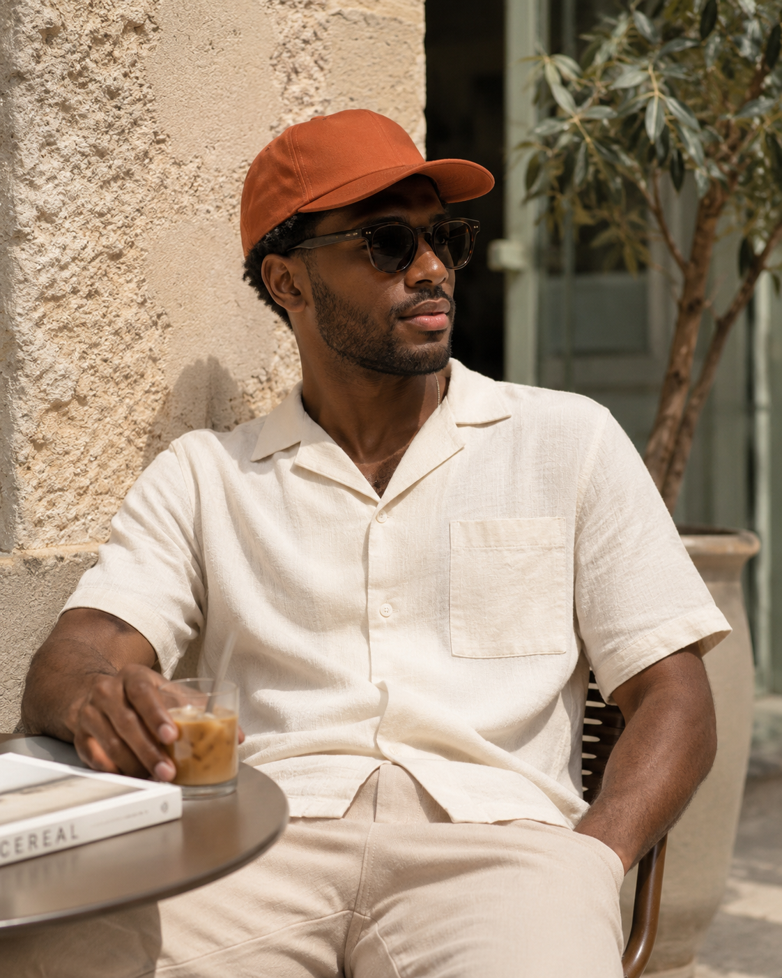 Man sitting outdoors wearing a beige shirt, orange cap, and sunglasses, holding a drink.