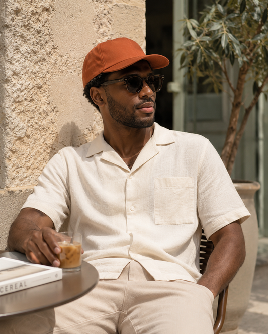 Man sitting outdoors wearing a beige shirt, orange cap, and sunglasses, holding a drink.