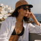Woman wearing a white shirt over a black bikini top with sunglasses and a cap, sitting by the ocean.