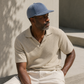 Man wearing a blue cap and beige shirt sitting against a light-colored wall.