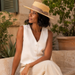 Woman in a white dress and straw hat sitting outdoors with plants in the background