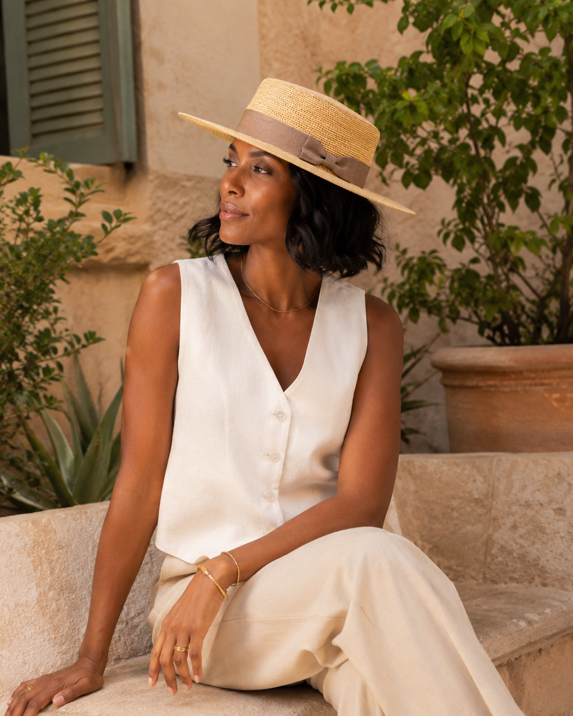 Woman in a white dress and straw hat sitting outdoors with plants in the background