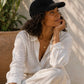 Woman wearing a black cap and white shirt sitting outdoors with plants in the background