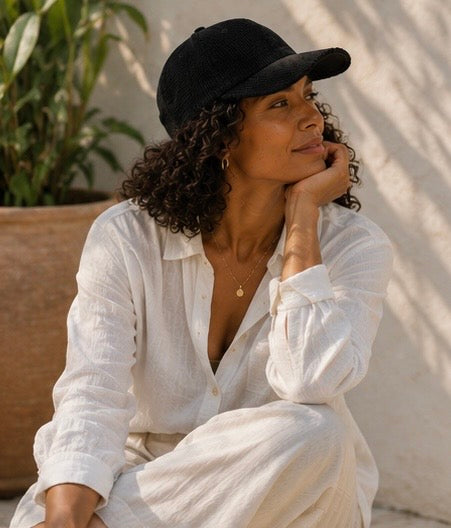 Woman wearing a black cap and white shirt sitting outdoors with plants in the background