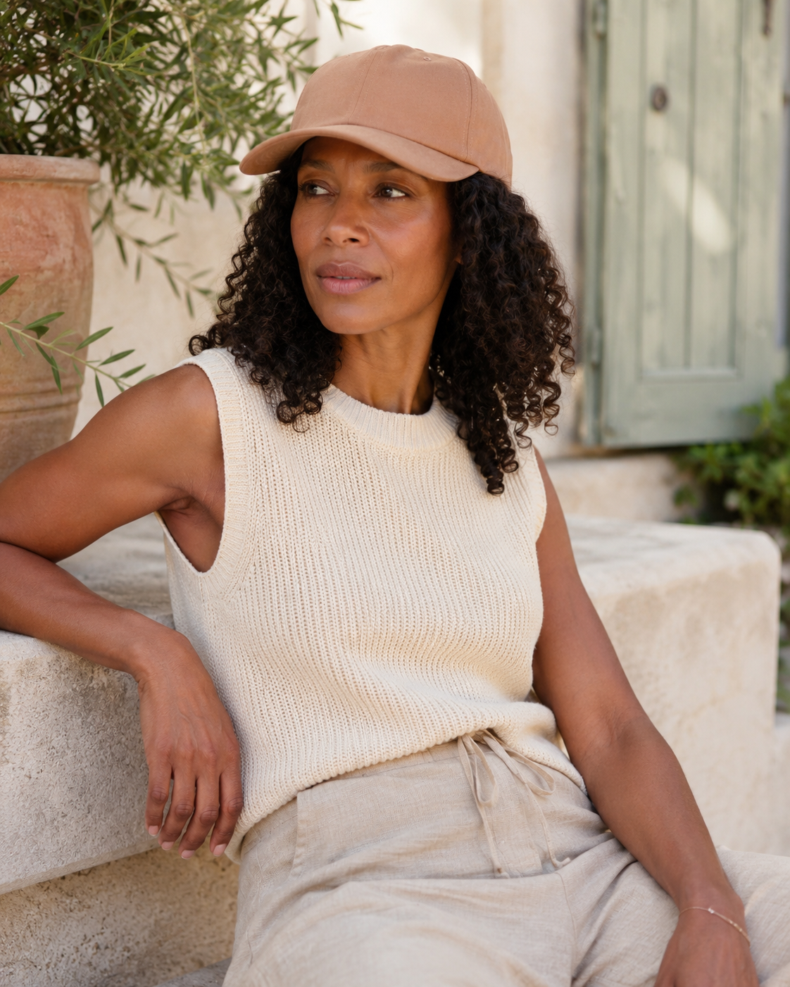 Woman wearing a beige cap and sleeveless top, sitting outdoors.