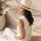 Woman wearing a white hat and dress sitting in a sunlit room with plants.
