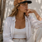 Woman wearing a white shirt, beige pants, and a brown cap sitting outdoors.