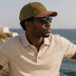 Man wearing a beige polo shirt and green cap with sunglasses, standing by the ocean.