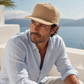 Man wearing a beige cap and light blue shirt sitting outdoors with a scenic background