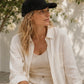 Woman wearing a black cap and white outfit sitting outdoors with greenery in the background