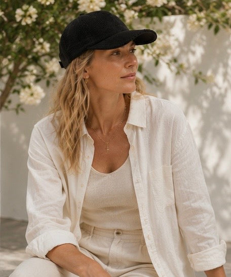 Woman wearing a black cap and white outfit sitting outdoors with greenery in the background