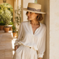 Woman wearing a white shirt and beige hat leaning against a wall with plants in the background
