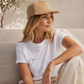 Woman wearing a beige cap and white t-shirt sitting on a light-colored surface with a neutral background