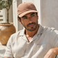 Man wearing a brown cap and light-colored shirt sitting outdoors.