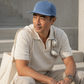 Man sitting outdoors wearing a blue cap and beige outfit with a white bag.