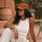 Woman wearing a white sleeveless top and beige pants sitting outdoors with a terracotta cap.