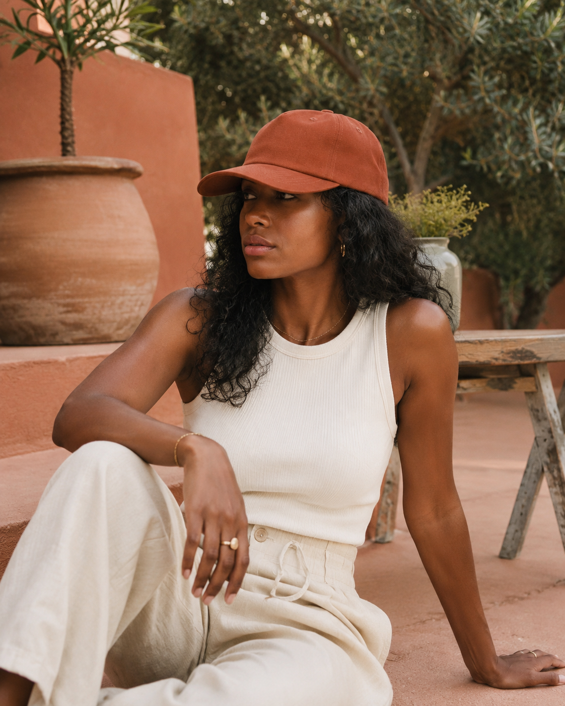 Woman wearing a white sleeveless top and beige pants sitting outdoors with a terracotta cap.